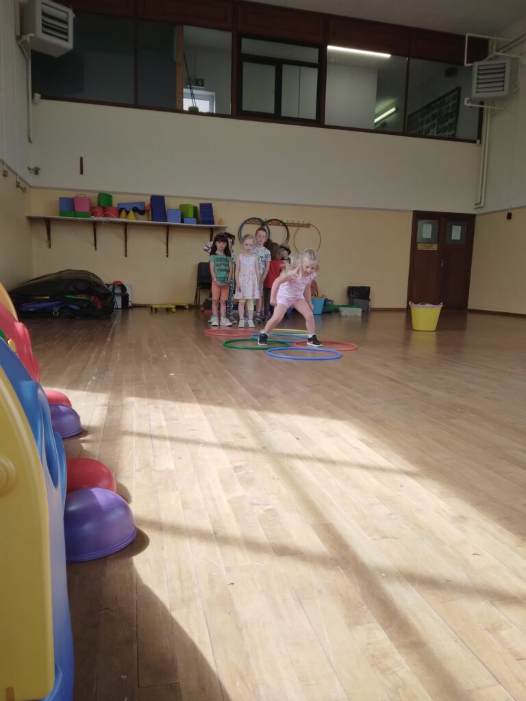 Children jumping into coloured hoops in a wooden floored hall. 