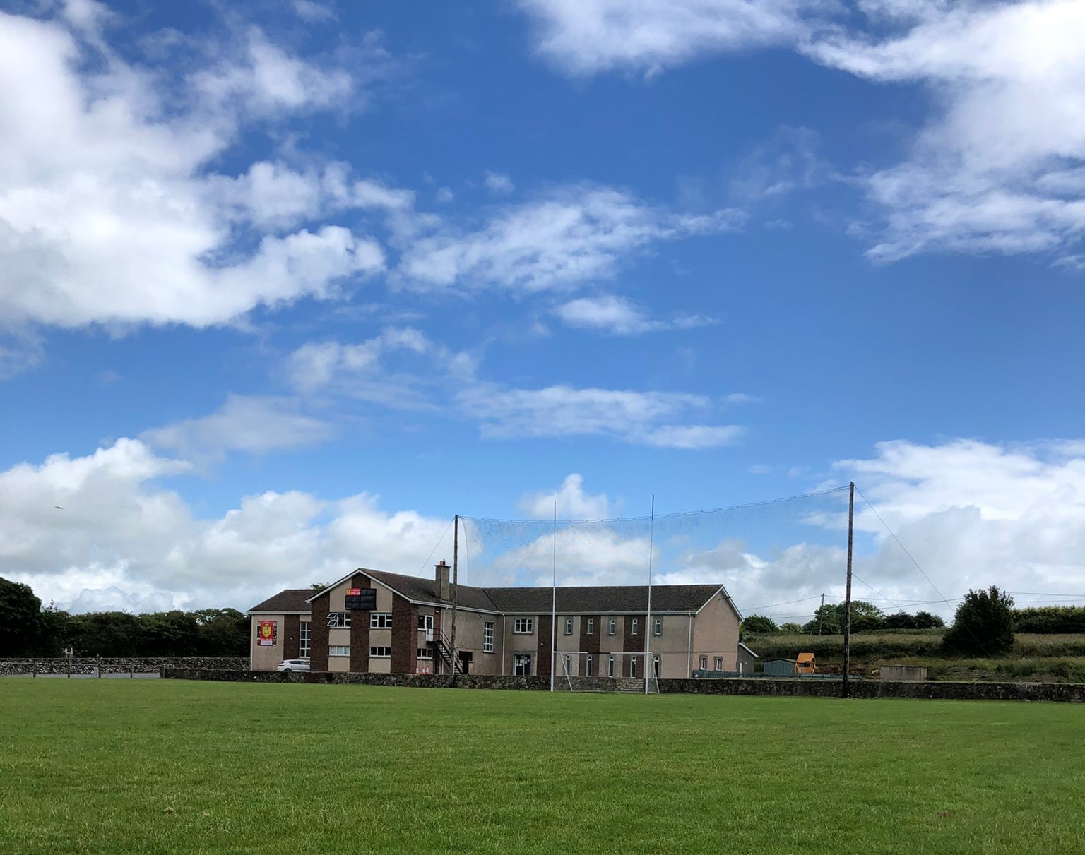 image of Dunhill Sports Centre from the middle of a sports pitch. The building is in the centre of the picture, grass to the front, blue sky and clouds over the building and hedgerow to the right of the building