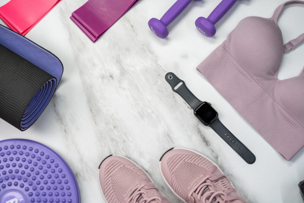 Mixture of gym items. pink runners, black watch, pink sports bra, purple hand weights, dark pink stretch band, rolled up black mat, purple disc